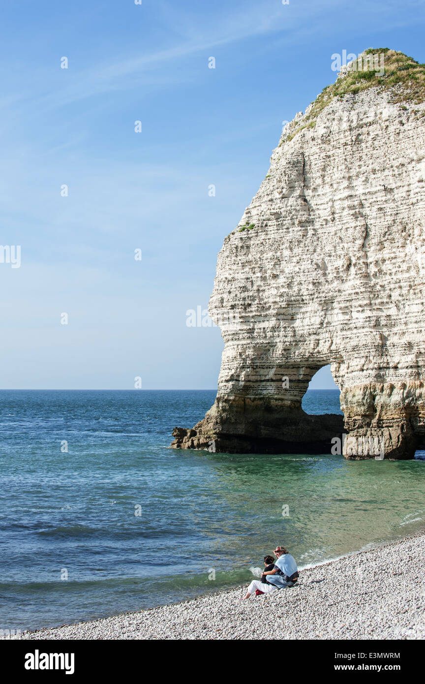 Couple sitting on pebble beach in front of the natural rock arch porte d'Amont at Etretat, Normandy, France Stock Photo