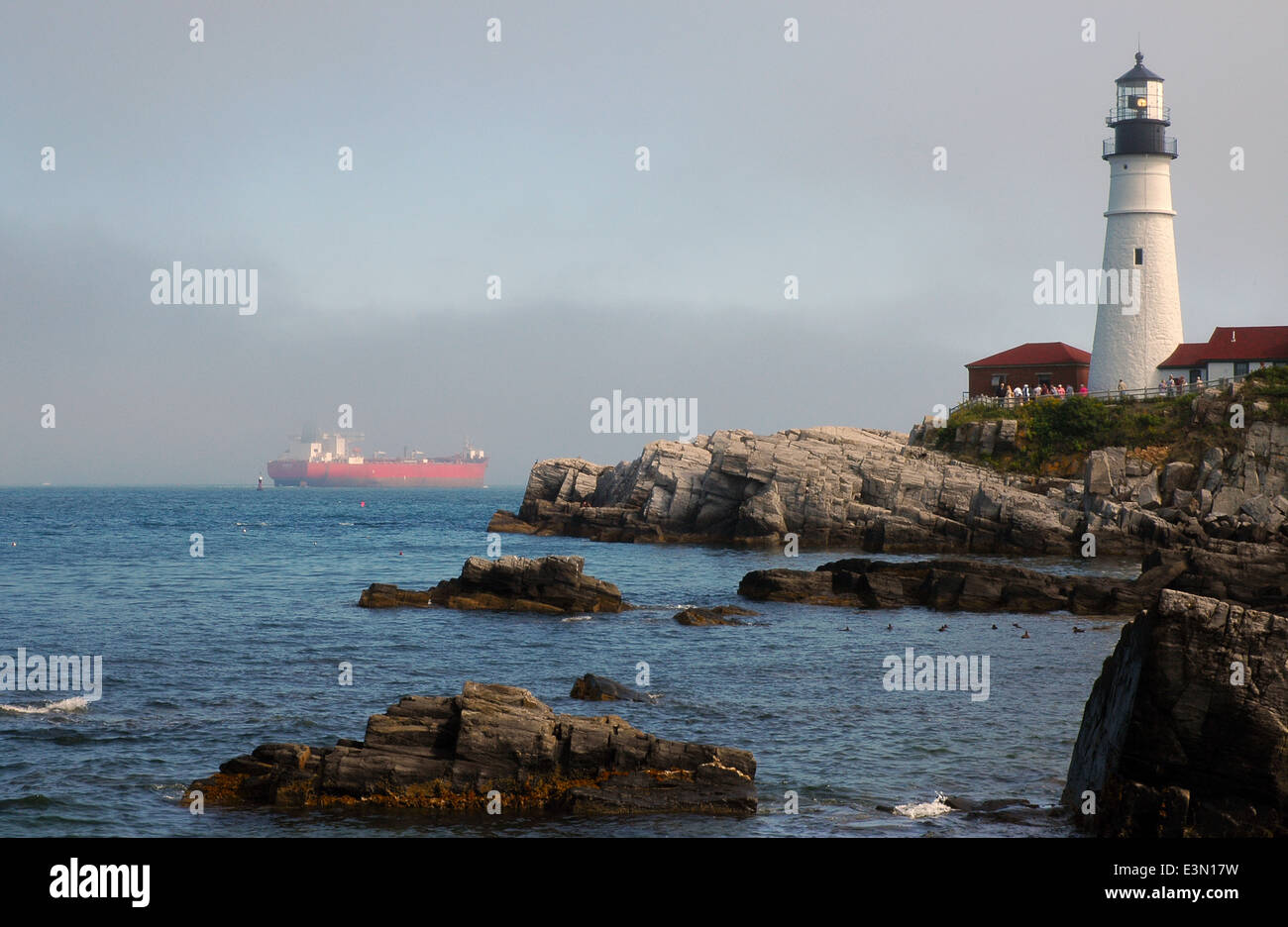 Portland Head lighthouse guiding red tanker ship through fog along the rocky coast. It has been guiding mariners since Colonial times. Stock Photo