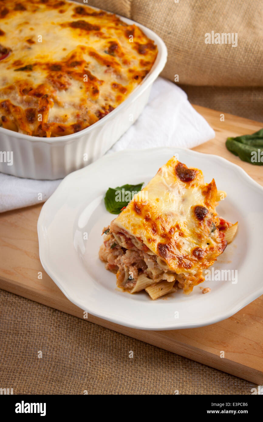 Italian baked ziti serving on plate with full casserole dish in background. Fork with bite on it. Looks like lasagna Stock Photo