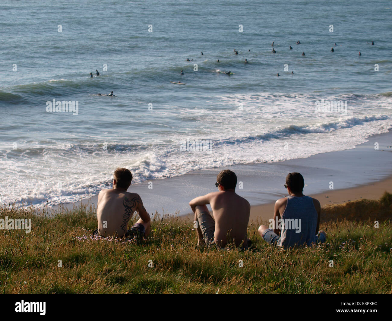 Three young men sat on the cliff top looking out to sea, Widemouth Bay, Bude, Cornwall, UK Stock Photo