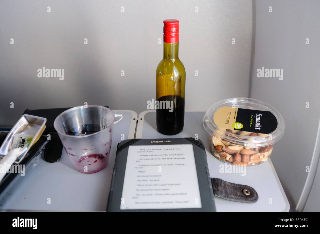 A half drunk bottle of red wine, a bowl of nuts and a Kindle on a seat table on board an airplane Stock Photo