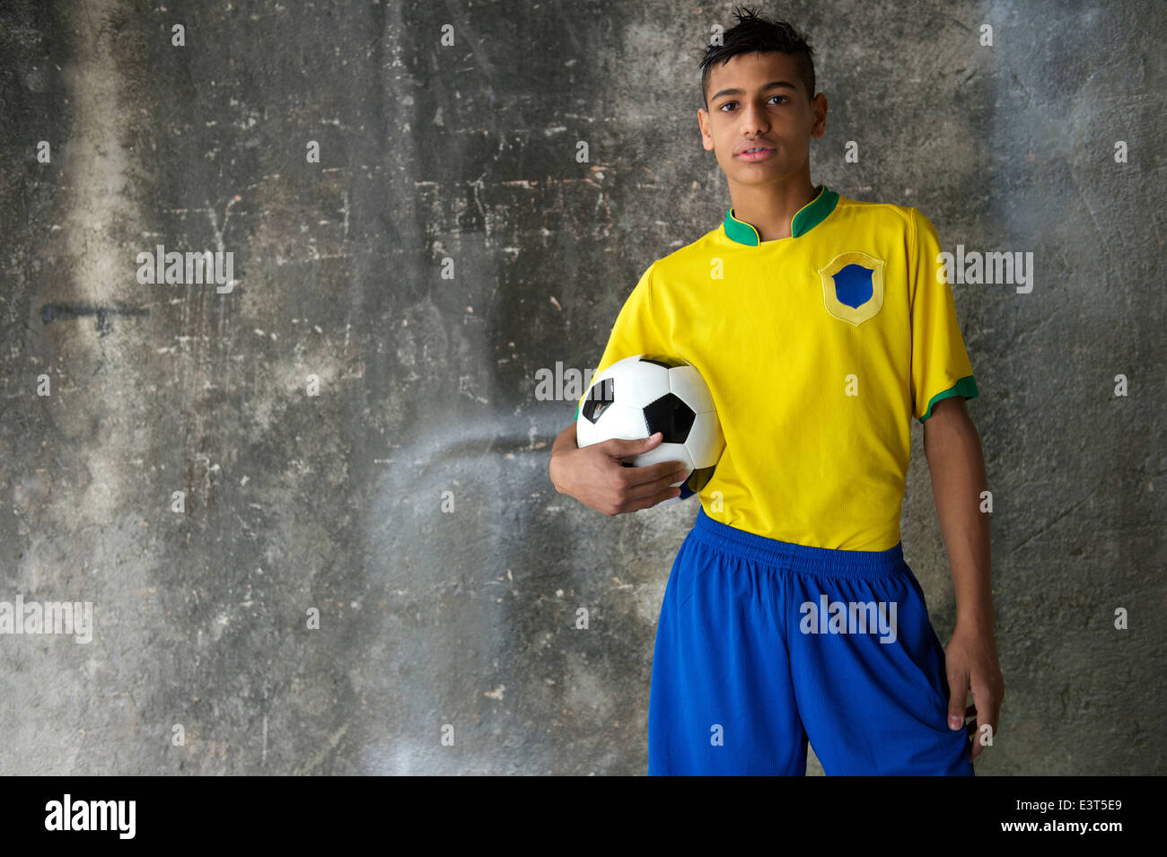 Young Brazilian footballer in team Brazil colors kit holding soccer ball against concrete favela wall Stock Photo