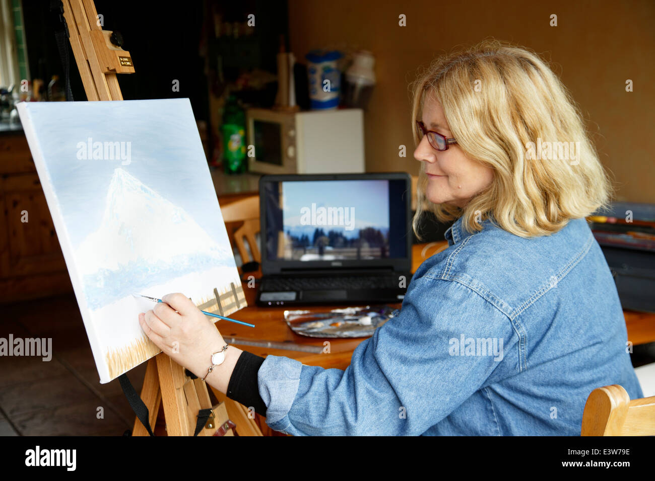 Woman painting in kitchen using photograph on computer screen as reference Stock Photo