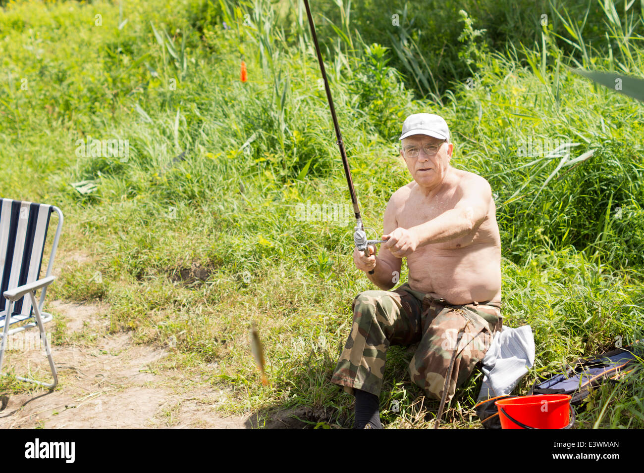 Elderly disabled fisherman sitting on a lake shore reeling in a small fish on his rod and line Stock Photo