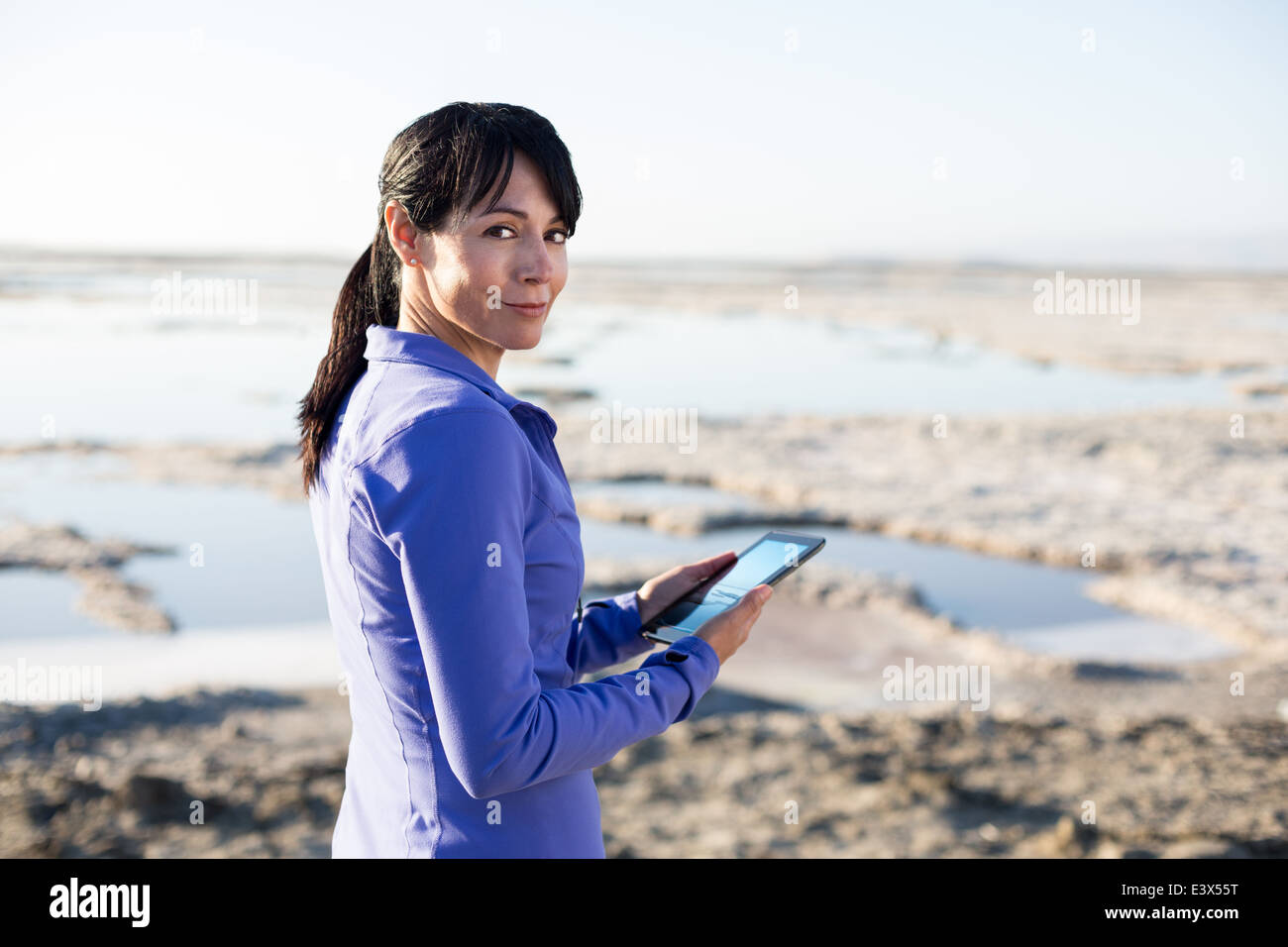 Sporty fortysomething woman in purple smiling over her shoulder using smalll tablet device as a camera outdoors. Stock Photo