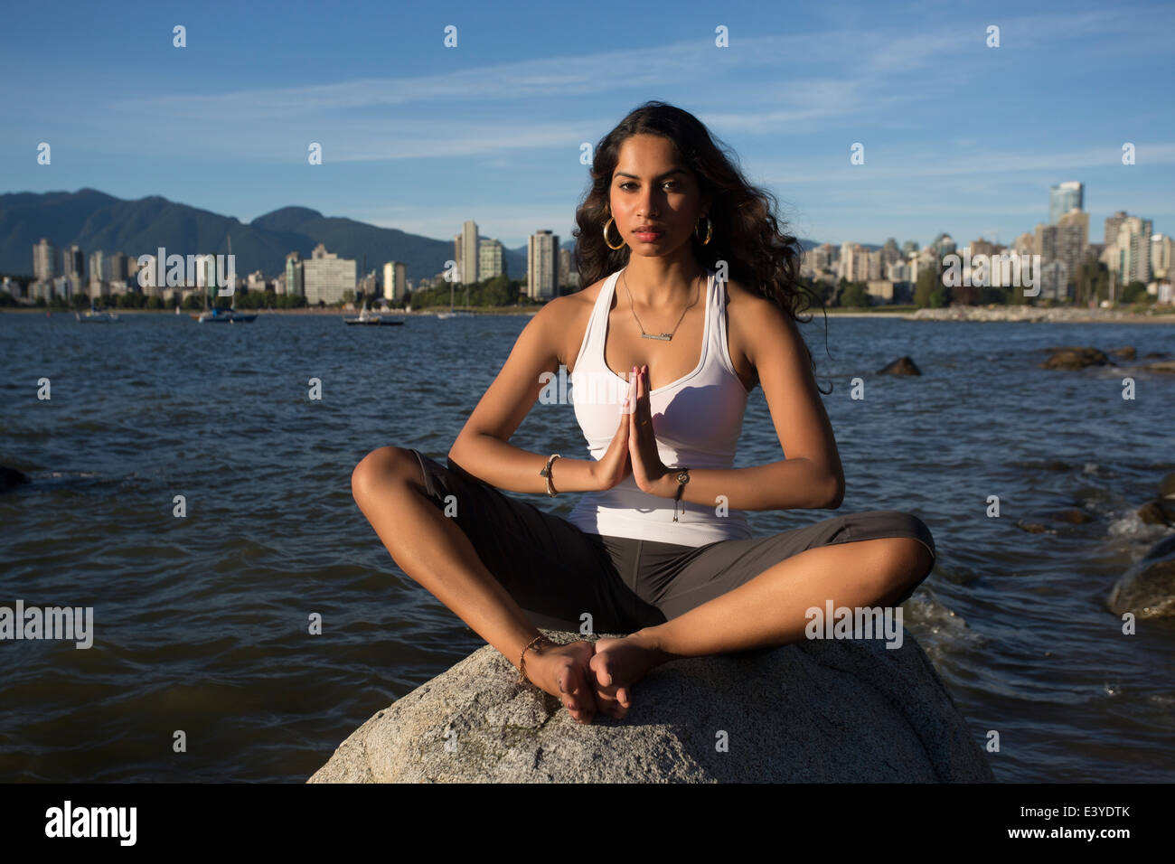 A young Indian woman is meditating on a rock with the ocean and downtown Vancouver in the distant background. Stock Photo