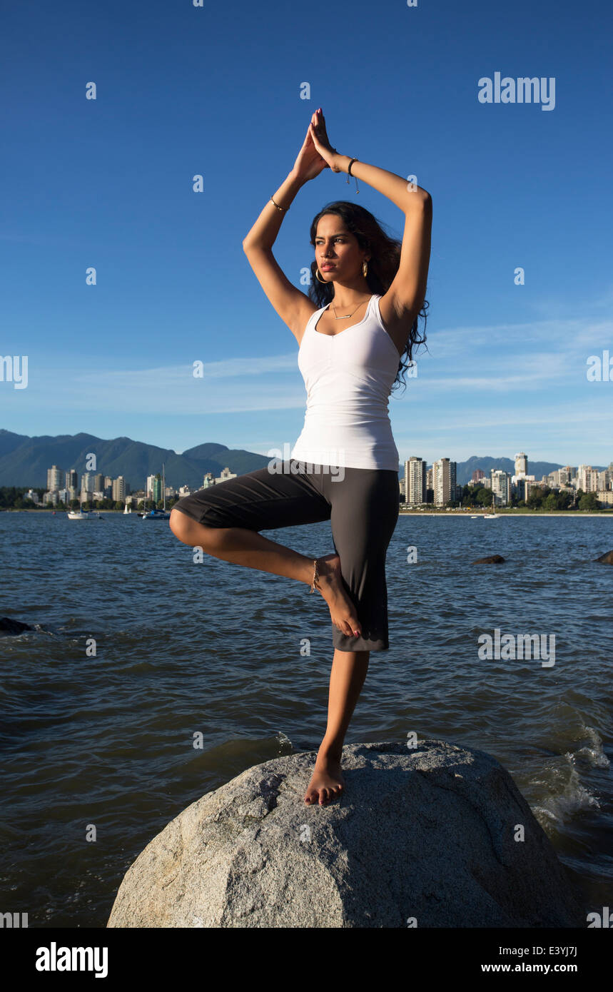 Young Indian woman doing yoga (standing on one leg) on a rock with the city skyline of Vancouver in the distant background. Stock Photo