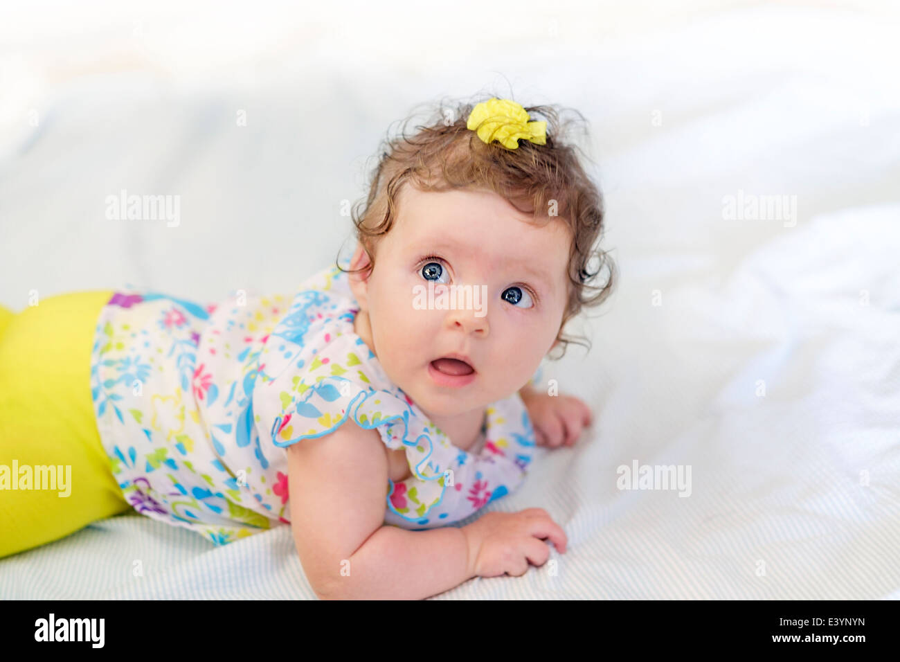 A three month old Caucasian infant girl, wearing yellow and lying on stomach on a white blanket and looking up. Stock Photo