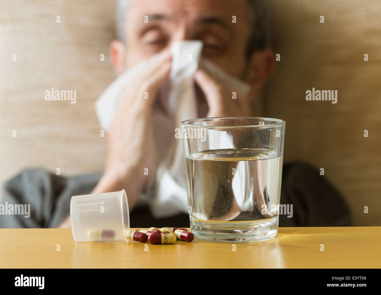 Glass of water and 500mg Amoxicillin antibiotic tablets on table next to man with cold blowing his nose. Stock Photo