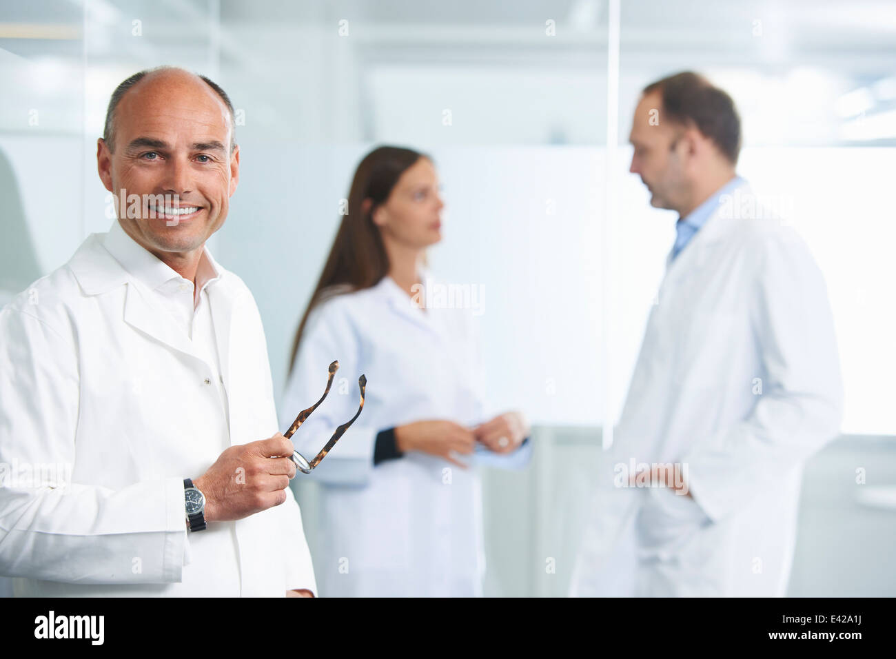 Male doctor by reflective wall, colleagues in background Stock Photo