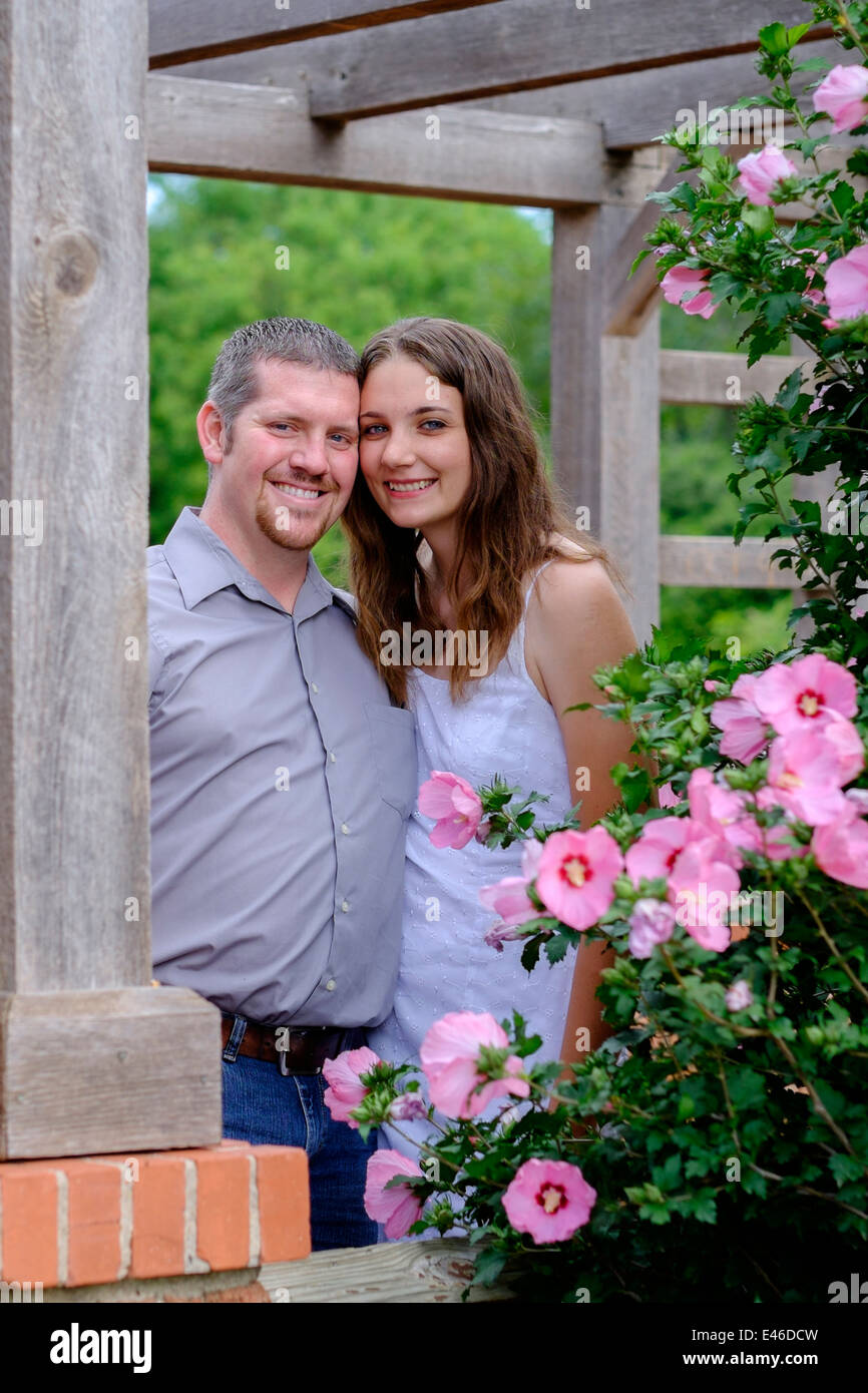 A concepts image of a happy, loving couple in their 20s pose for engagement pictures in a beautiful park surrounded by colorful blooming flowers. USA. Stock Photo