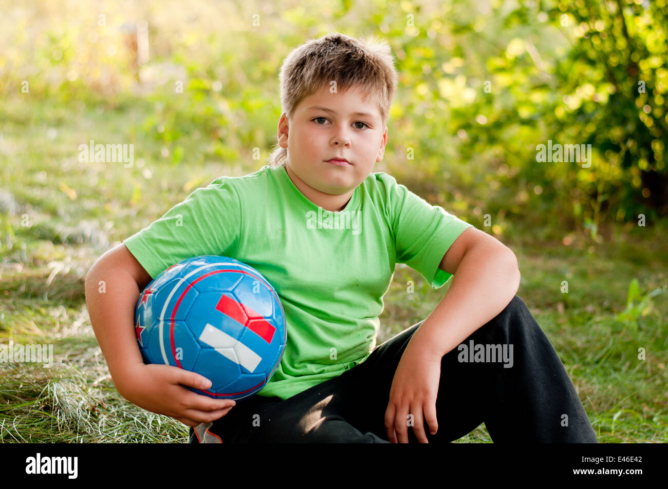 boy child alone ball soccer football eight years nature park square summer day sunny sports t-shirt grass sport blond sitting ma Stock Photo