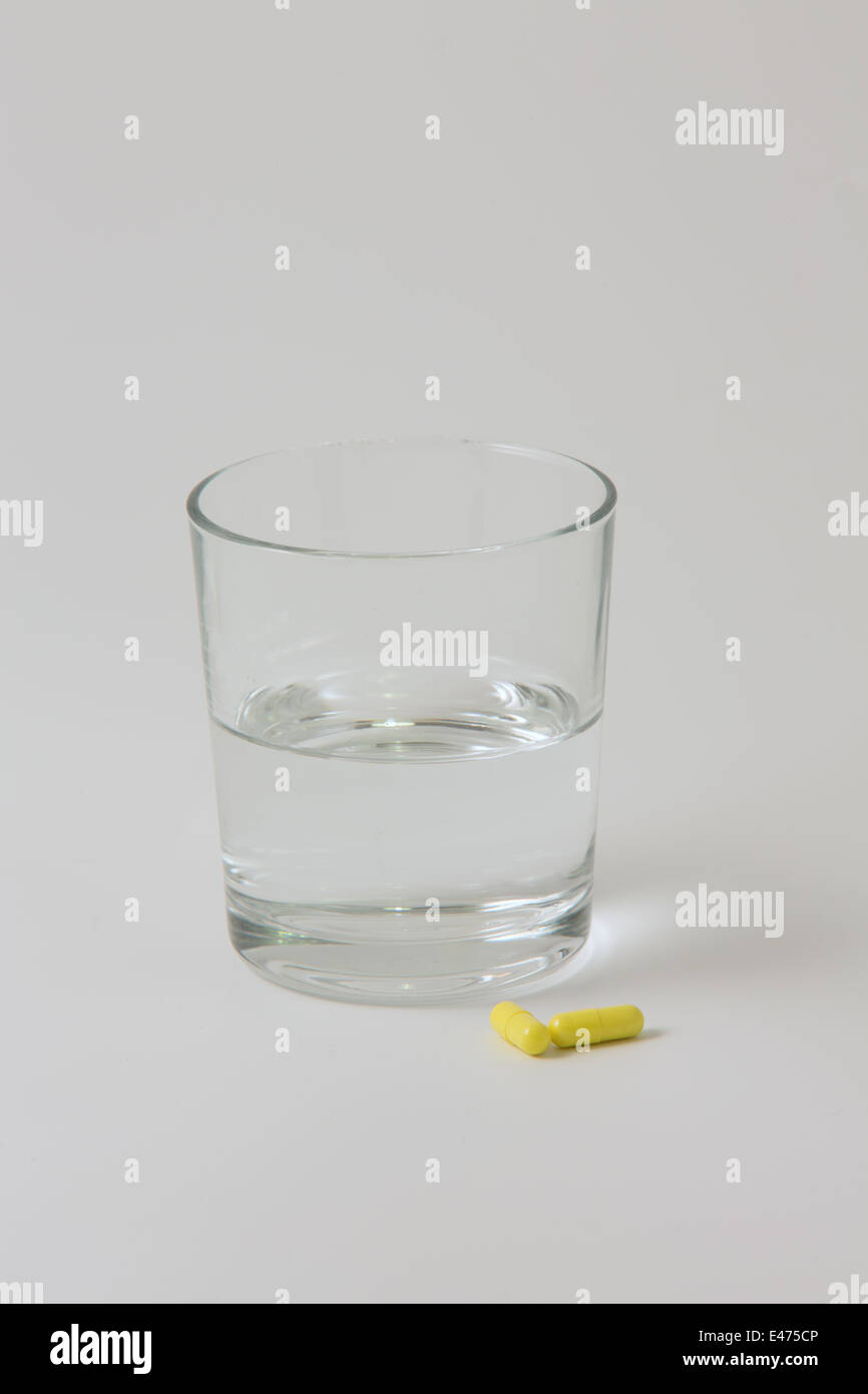 Two yellow tablets next to a small, clear glass of water with a plain white background Stock Photo