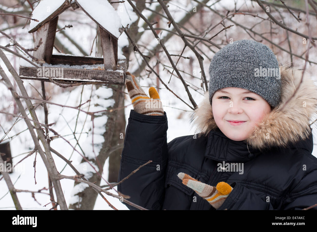 boy child one winter feeding 9 8 years snow snow-covered jacket hat cap feed house garden park forest hanging Russia wood wooden Stock Photo