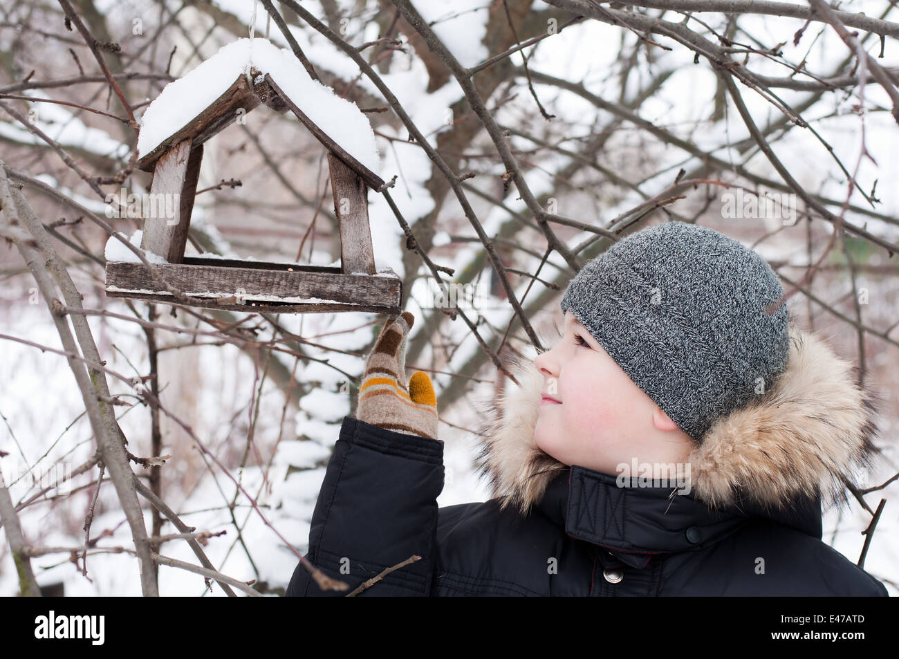 boy child one winter feeding 9 8 years snow snow-covered jacket hat cap feed house garden park forest hanging Russia wood wooden Stock Photo