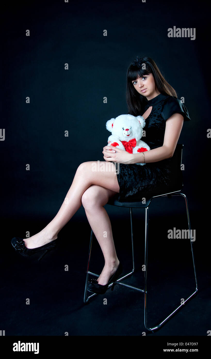 Beautiful european teenage girl is sitting on high chair, wearing short black dress, holding white toy bear Stock Photo