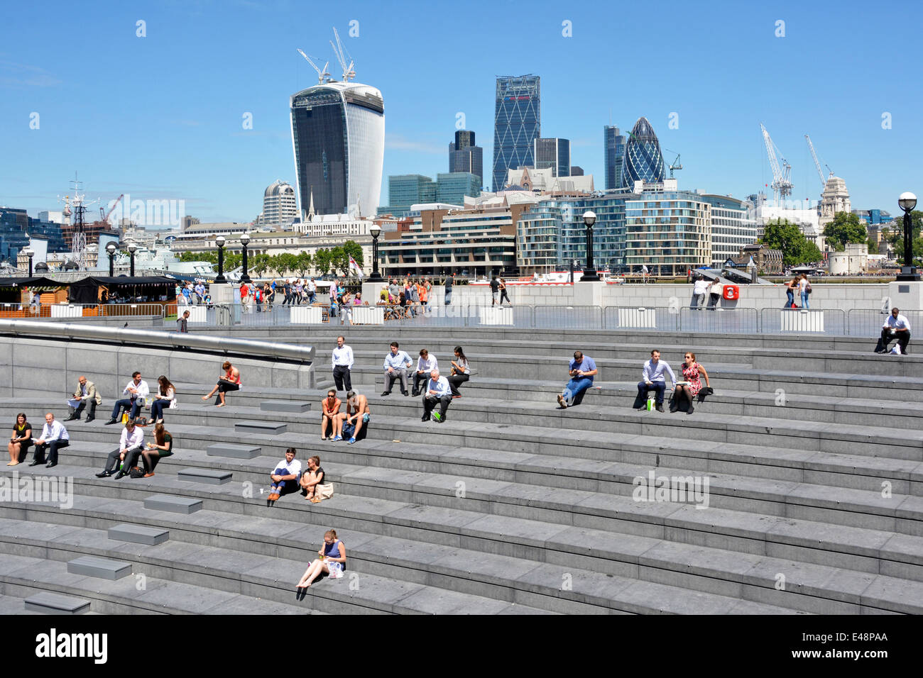 Blue sky sunny summer day tourists & office workers sitting in Scoop area at More London City landmark skyscraper skyline beyond Southwark England UK Stock Photo