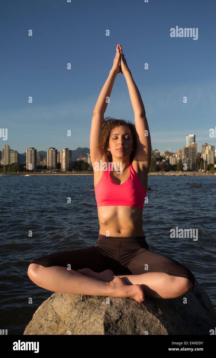 An attractive young woman wearing a pink top does yoga on a rock with the ocean and Vancouver background. Stock Photo
