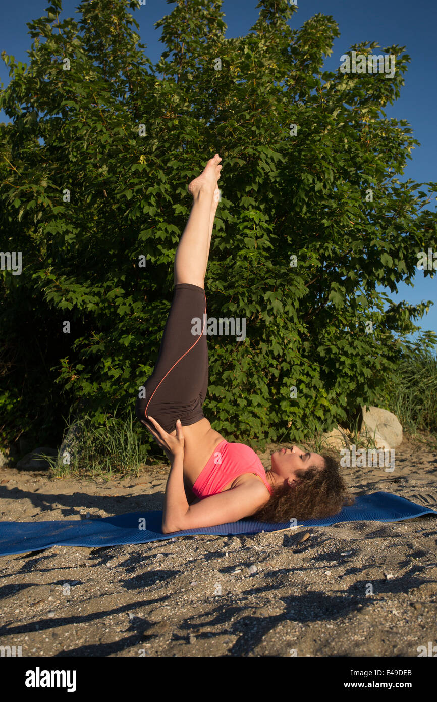 An attractive young woman does a shoulder stand on the beach wearing a pink top. Stock Photo