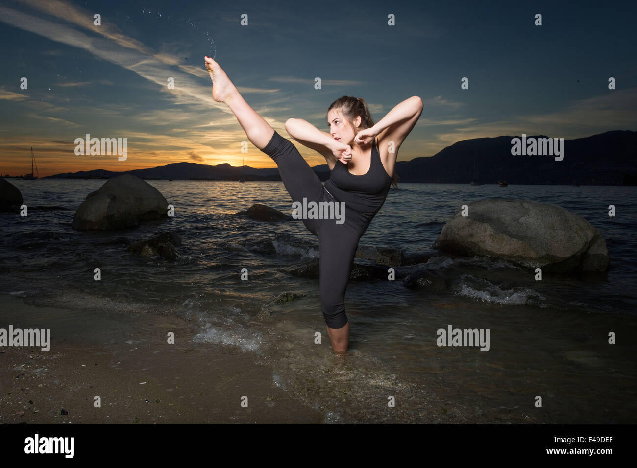A young woman performs a high gymnastic kick at the beach at sunset. Stock Photo