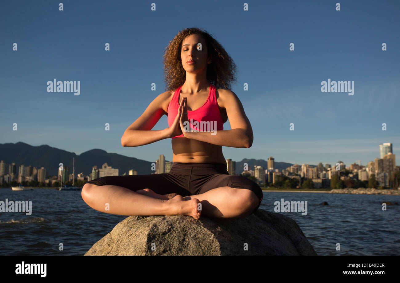 An attractive young woman wearing a pink top is meditating on top of a rock with the ocean and downtown Vancouver behind her. Stock Photo
