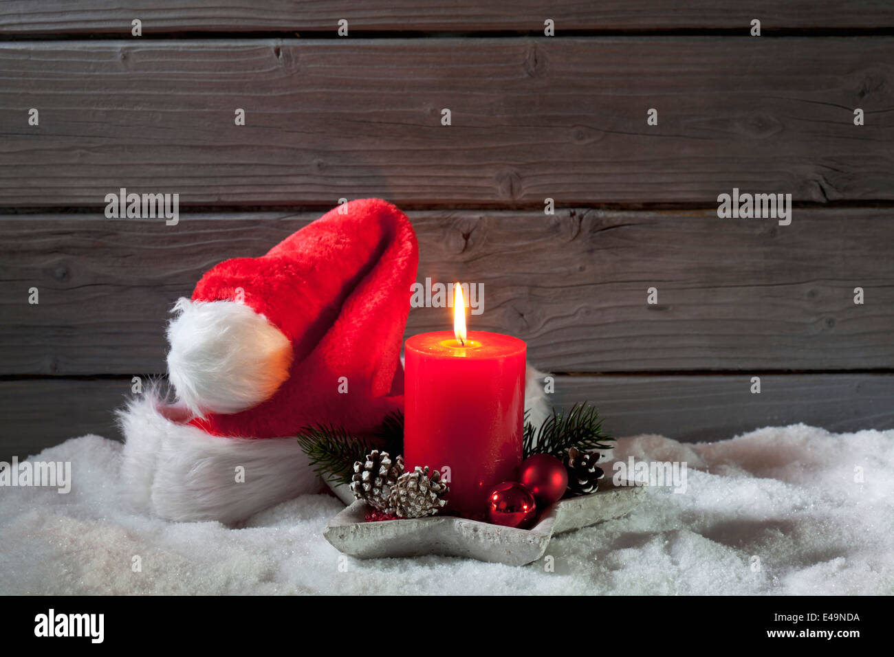 Lighted red candle and Christmas cap on artificial snow in front of grey wooden wall Stock Photo