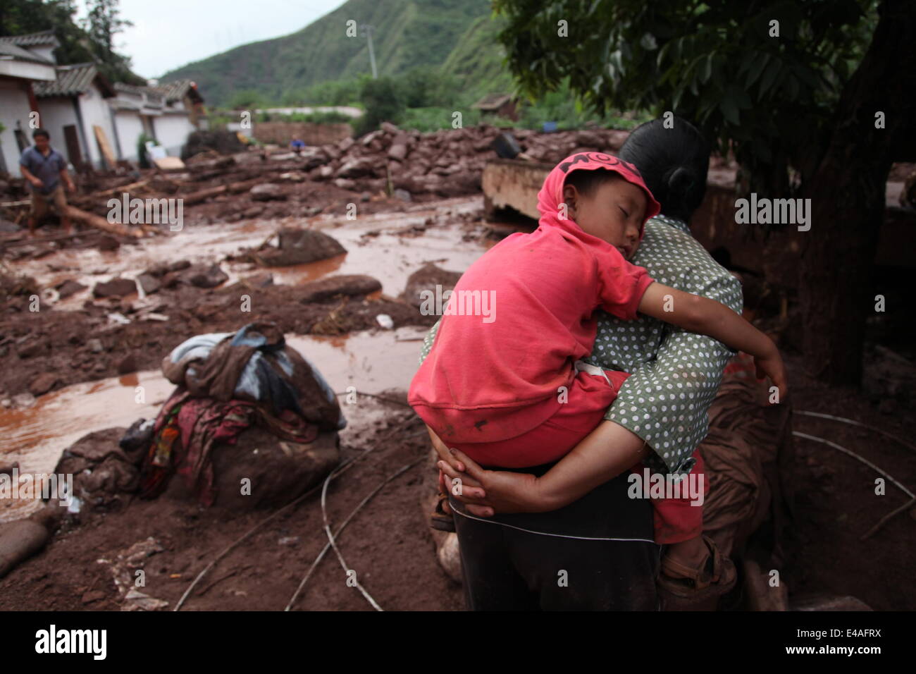 Zhaotong, China's Yunnan Province. 7th July, 2014. A woman carrying a child looks at damaged houses at mudslide-hit Longtoushan Town of Ludian County in Zhaotong City, southwest China's Yunnan Province, July 7, 2014. As of Sunday, continuous downpours battered Ludian County, leading to torrential floods and mudslides in multiple sites. By 4:00 p.m on Monday, four people were killed and seven others remained missing in rainstorm-triggered mudslides in worst-hit Longtoushan and Lehong townships. Credit:  Zhang Guangyu/Xinhua/Alamy Live News Stock Photo