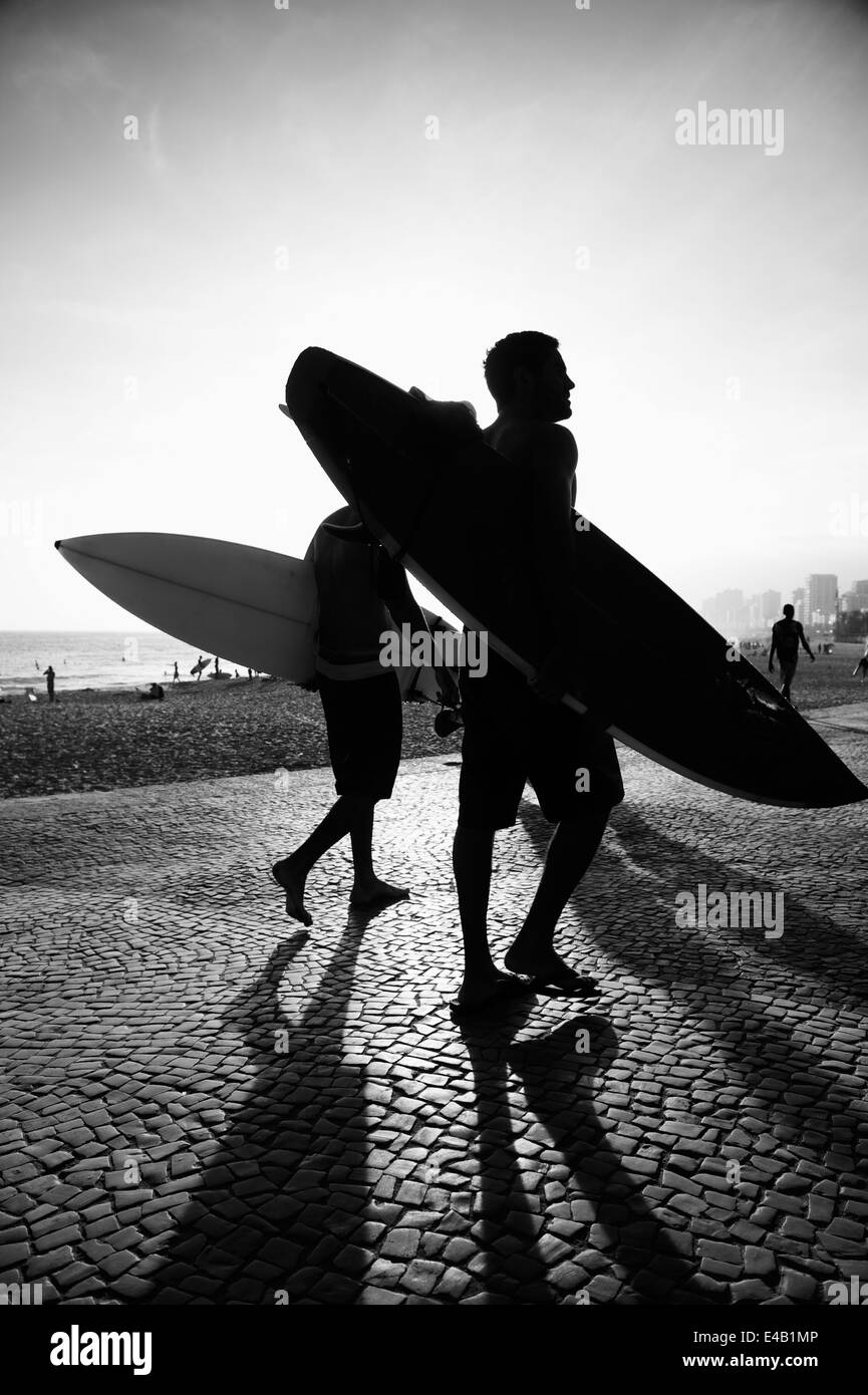 Sunset silhouettes of surfers walking at Arpoador Ipanema Beach Rio de Janeiro Brazil in black and white Stock Photo