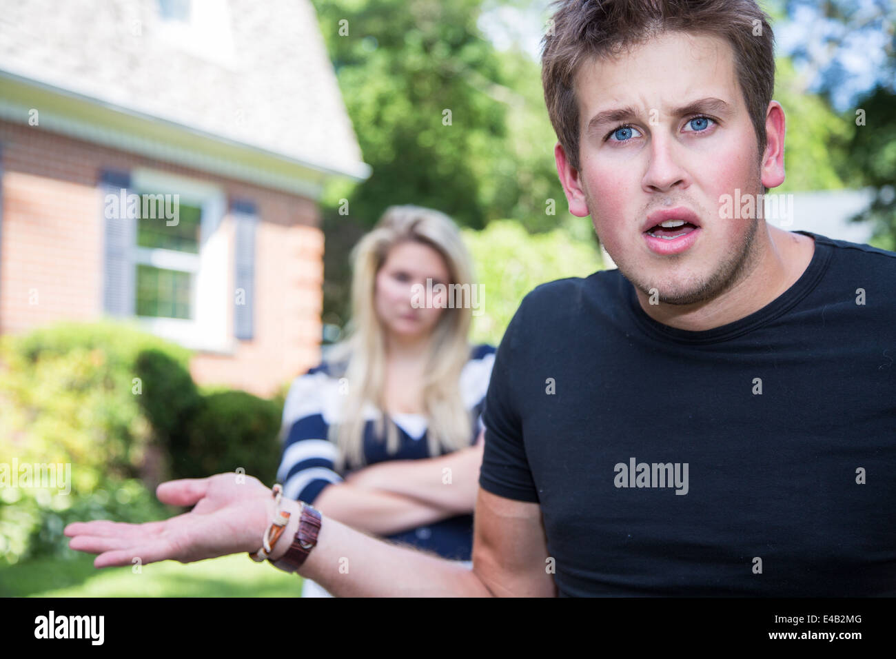 A bewildered and annoyed young man appears to feign innocence, while an angry young woman looks on from the background. Stock Photo