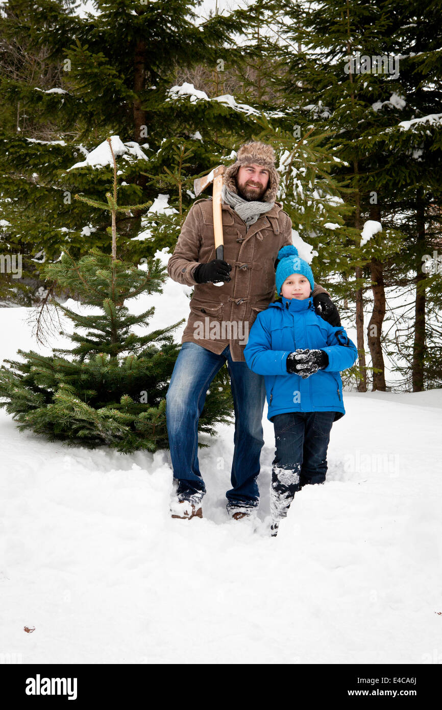 Father and son standing with axe by Christmas tree in snow-covered landscape, Bavaria, Germany Stock Photo