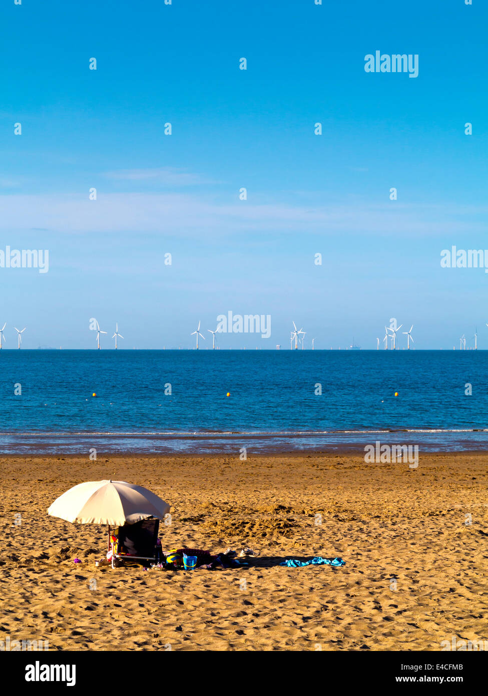 Person on deck chair underneath umbrella on beach at Colwyn Bay North Wales looking towards offshore wind farm in the Irish Sea Stock Photo