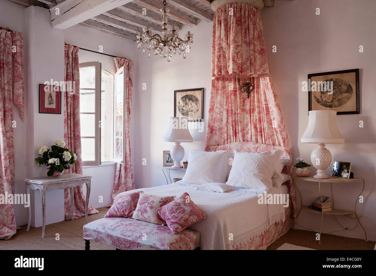 Pink and white toile de jouy fabric on cushions, curtain and bed coronet canopy in bedroom with wooden ceiling beams and bedside Stock Photo