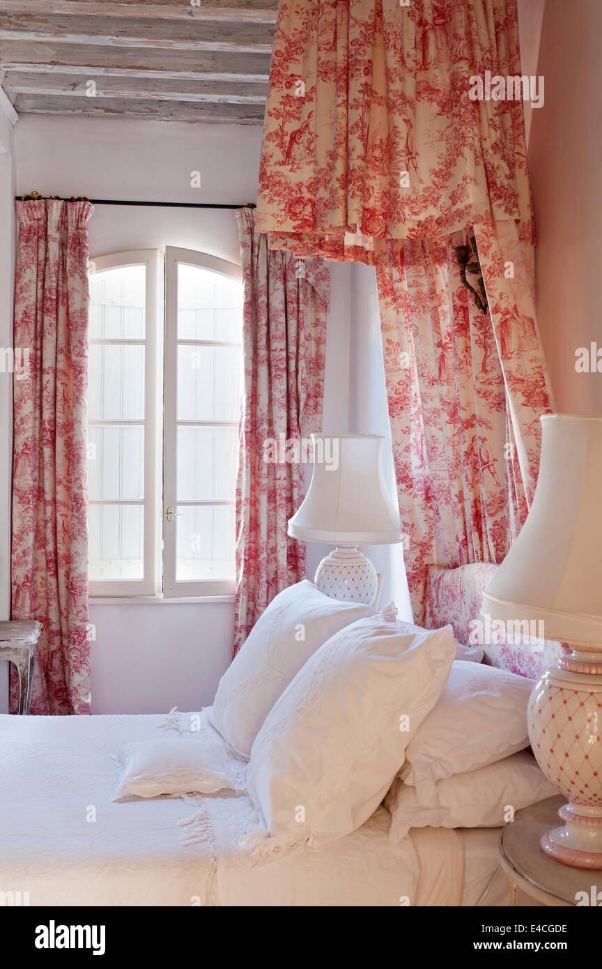 Pink and white toile de jouy fabric on curtains and bed coronet canopy in bedroom with wooden ceiling beams and bedside lamps Stock Photo