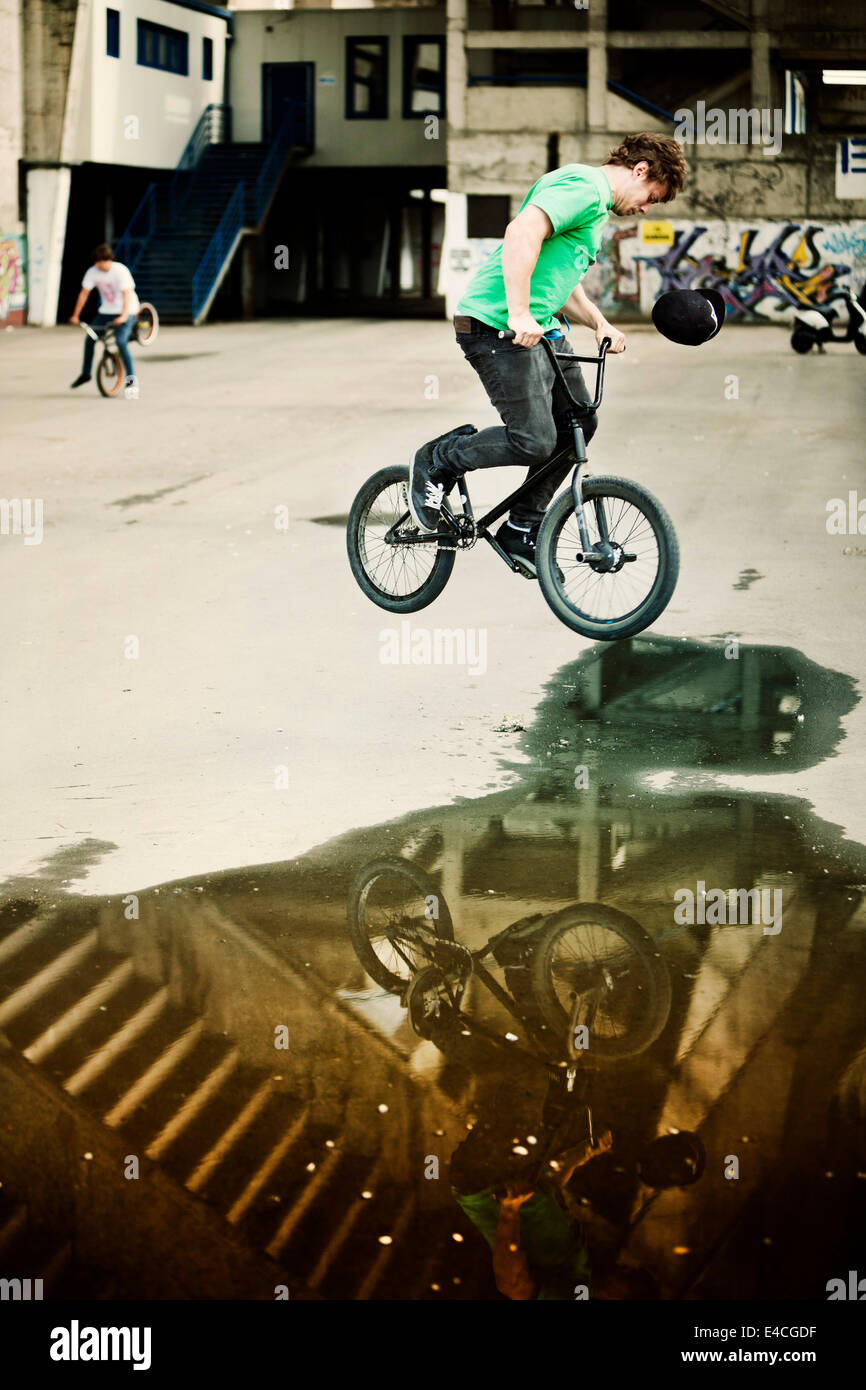 BMX biker performing a stunt over a puddle, man in background Stock Photo