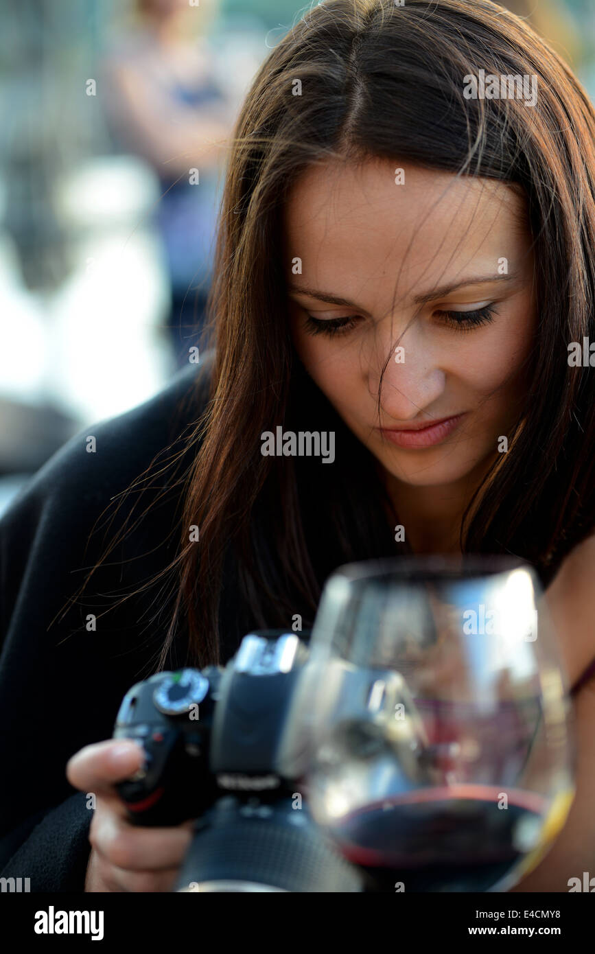 An attractive woman in her twenties viewing pictures on her camera screen while sitting having a glass of red wine Stock Photo