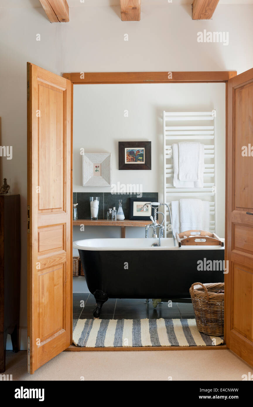 View through open double doors to bathroom with freestanding roll top bath and striped rug Stock Photo
