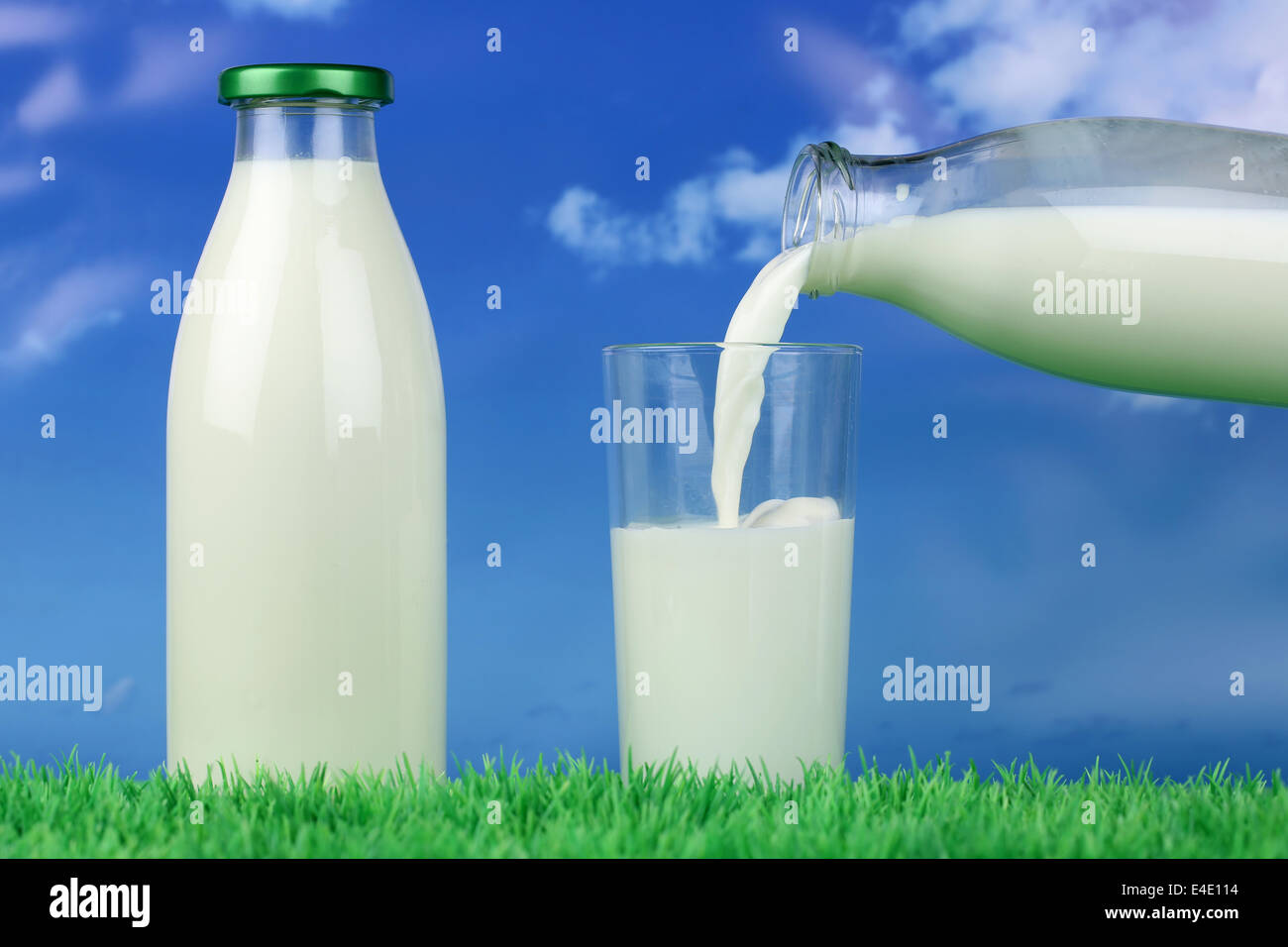 Fresh milk pouring from a bottle into a glass on a meadow in front of a blue sky Stock Photo