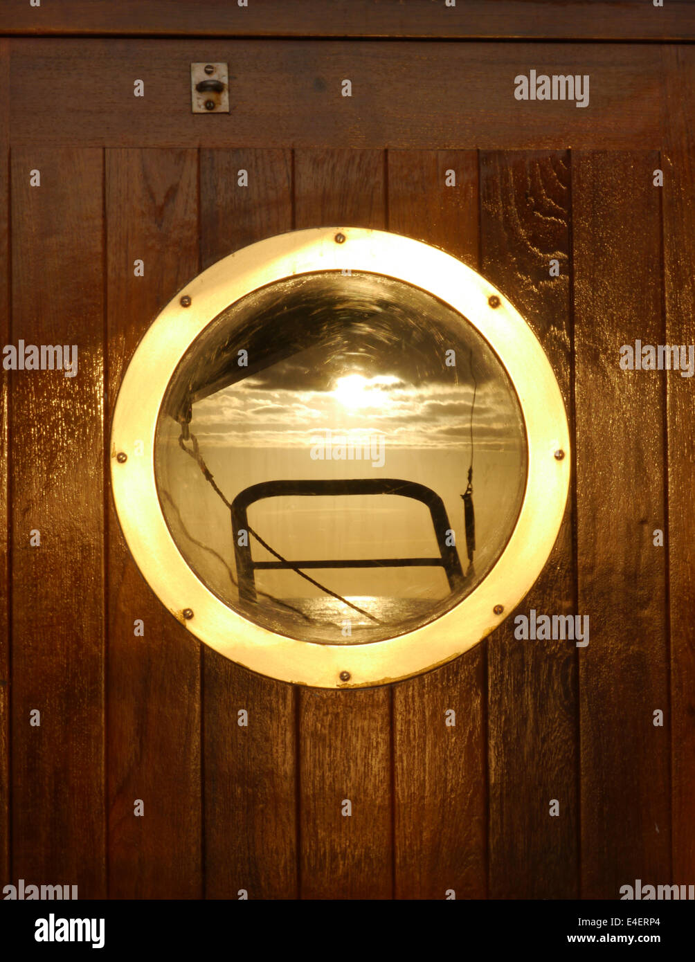 A close up of a varnished door on a ship with a brass porthole and a reflection of lifeboat harnesses Stock Photo
