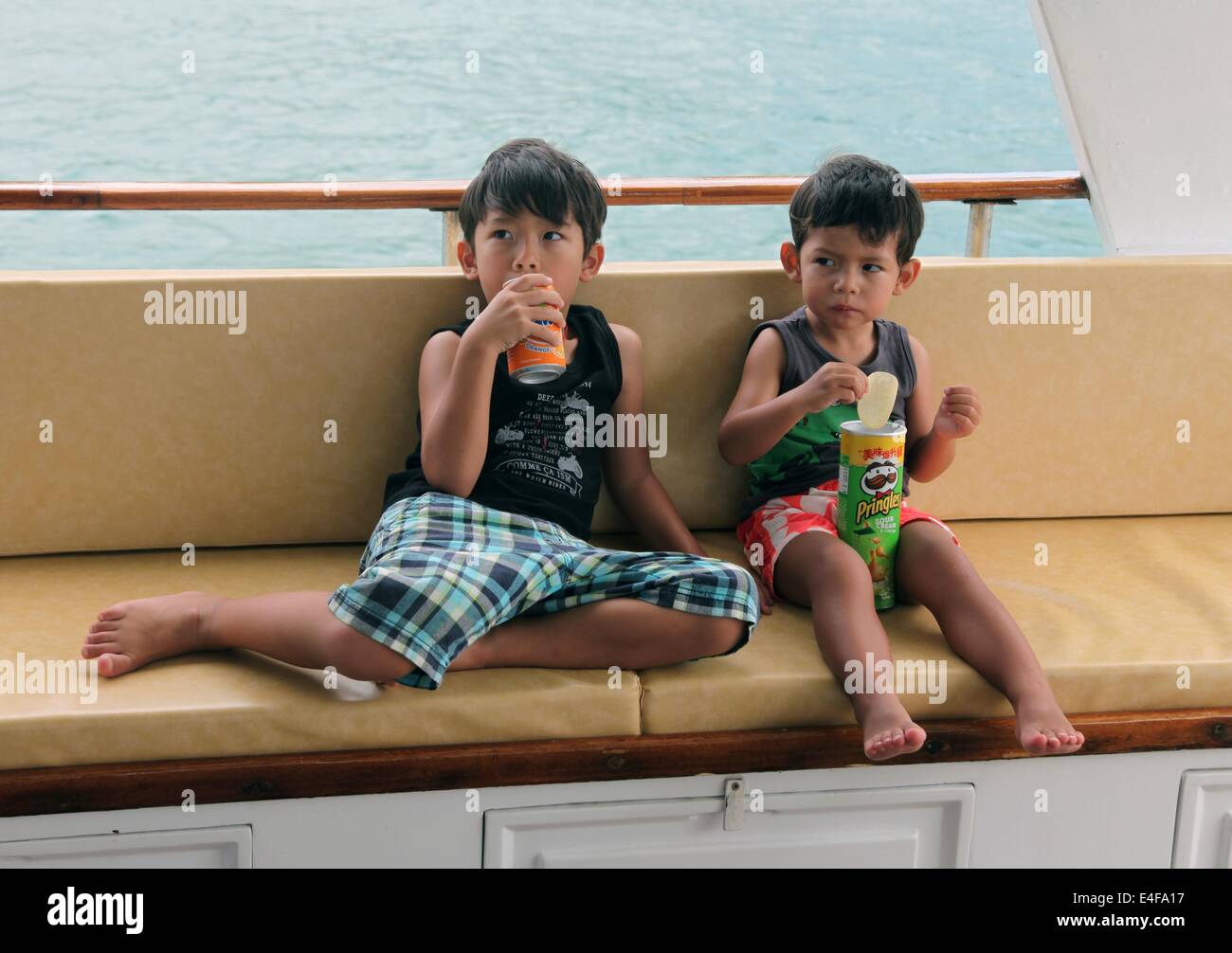 It's a photo of 2 child kid boys who sits on the deck of a boat or ship and eat and drink junk food like soda and chips Stock Photo