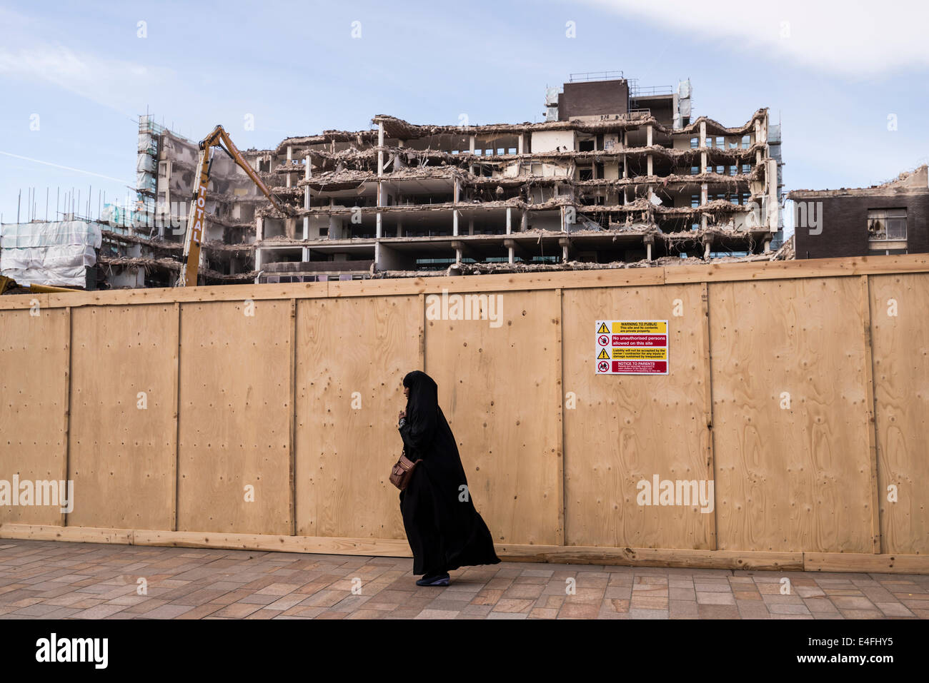 Building being demolished in background with woman in black Burqa walking past wooden fence Sheffield South Yorkshire England Stock Photo