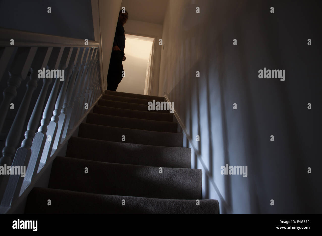 Silhouette of a man leaning against the wall at the top of a stairway, shadows cast on the walls from the light below. Stock Photo