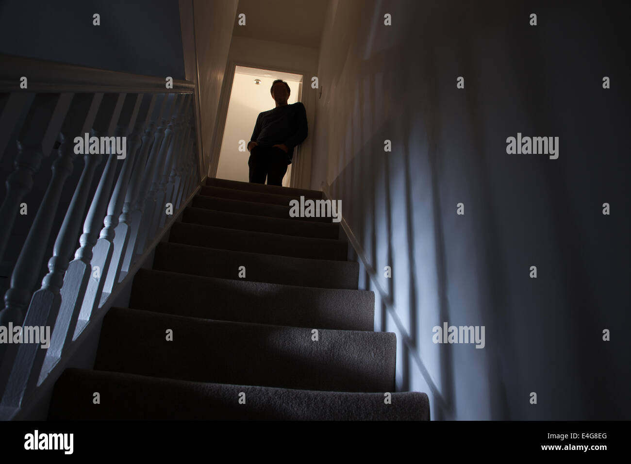 Silhouette of a man leaning against the wall at the top of a stairway, shadows cast on the walls from the light below. Stock Photo