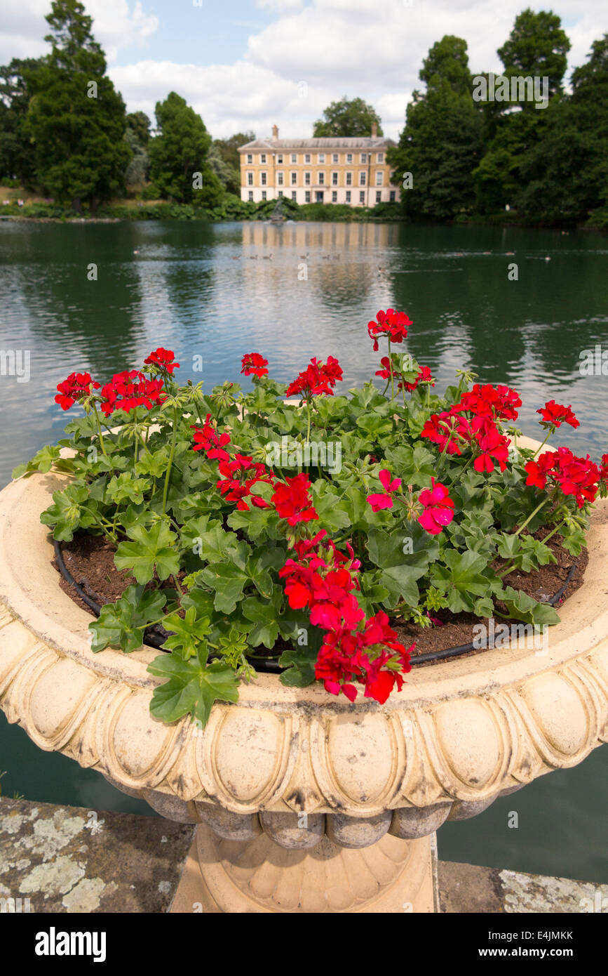 Red geraniums growing in Urn container pot with view towards Museum No 1 over Palm House Pond. Kew Gardens. Kew, West London UK. Stock Photo