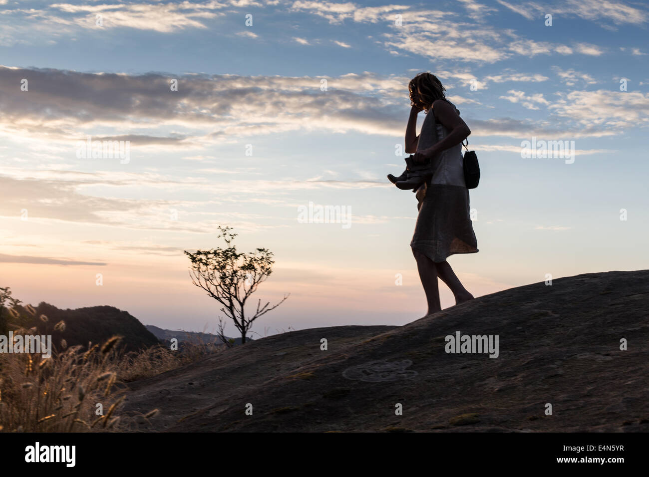 silhouette of woman on phone walking barefoot through remote hills at dusk holding shoes in hands Stock Photo