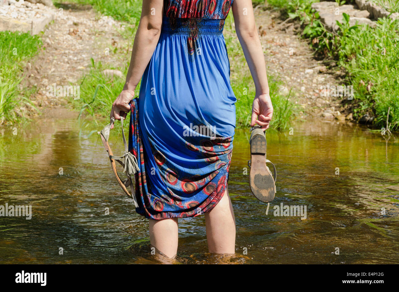 girl holds gray summer sandals and wade barefoot through the fast flowing stream in the park track Stock Photo
