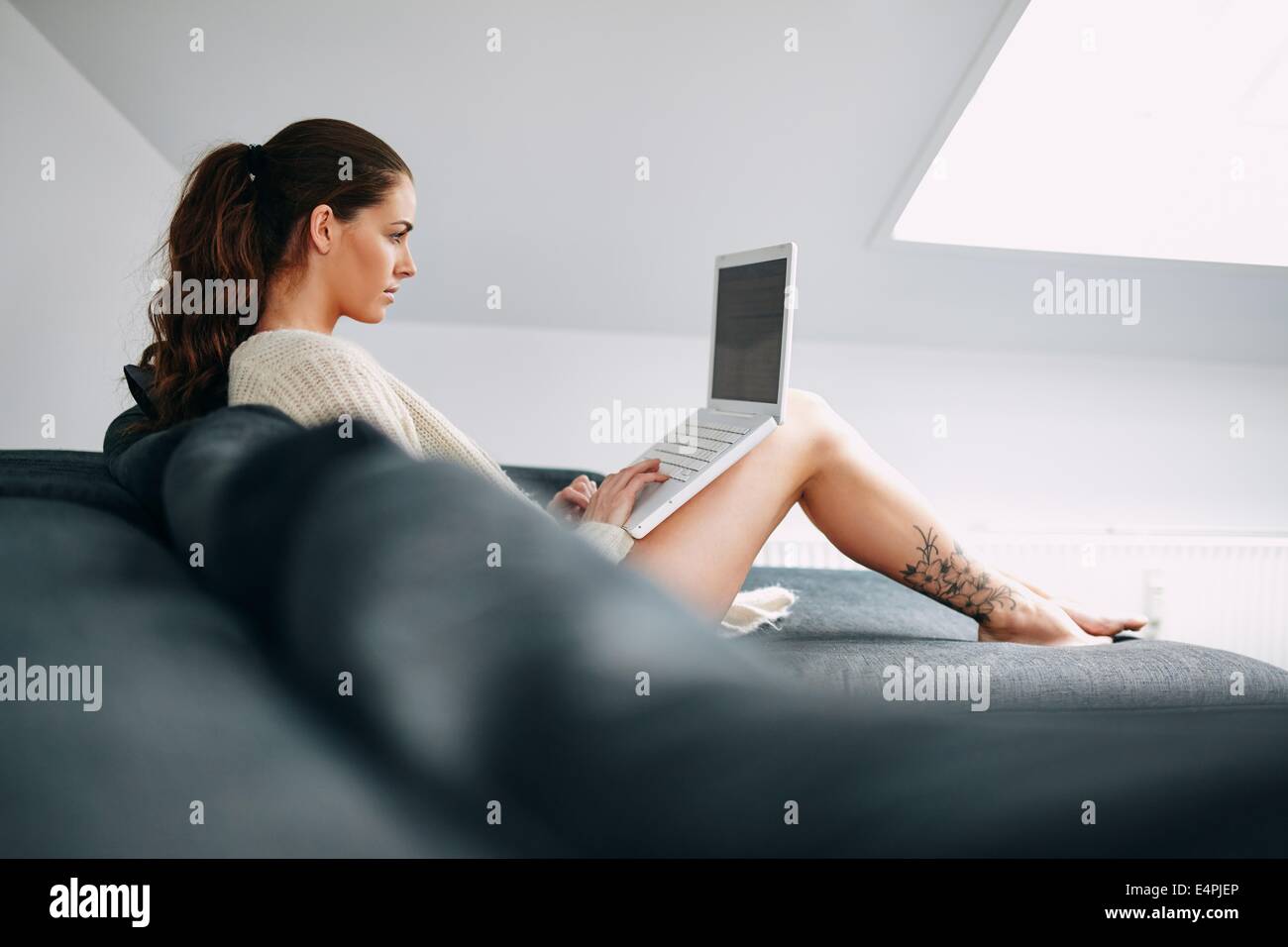 Image of pretty young brunette using laptop at home. Young female working on laptop while sitting on sofa. Stock Photo