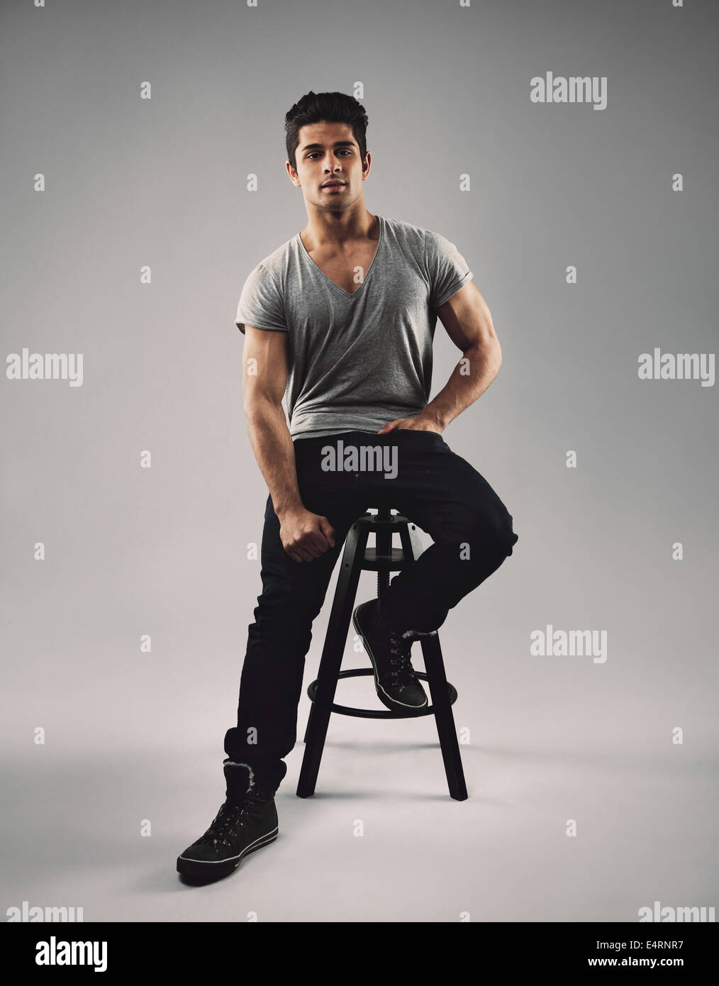 Full length portrait of handsome young man sitting on bar stool. Young hispanic male model over grey background. Stock Photo