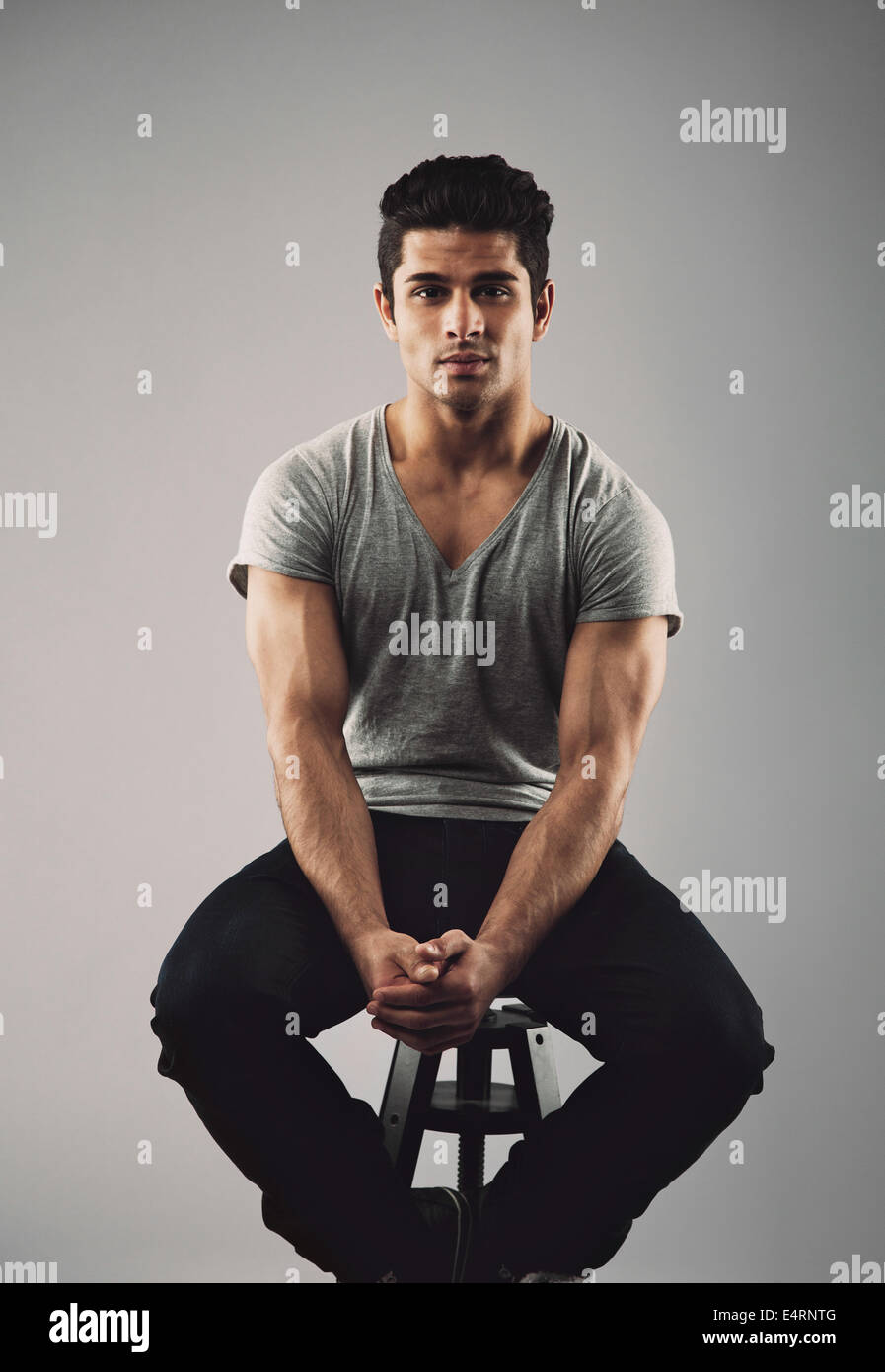 Portrait of muscular young male model sitting on stool. Hispanic young man over grey background. Stock Photo