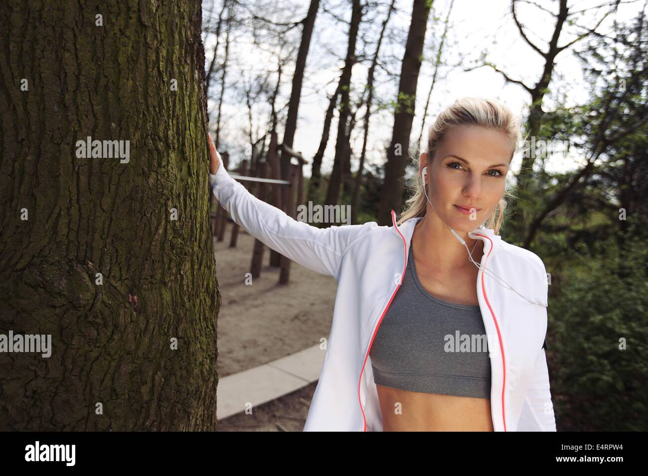 Pretty young woman standing by a tree at park. Attractive fitness female resting after outdoors training. Stock Photo