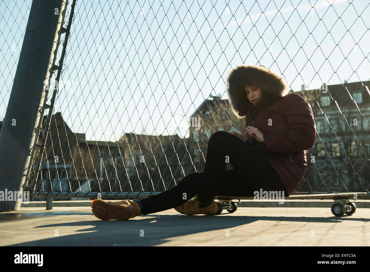 Teenage girl sitting on skateboard next to chain link fence near comercial dock, wearing winter coat, using smart phone, Germany Stock Photo